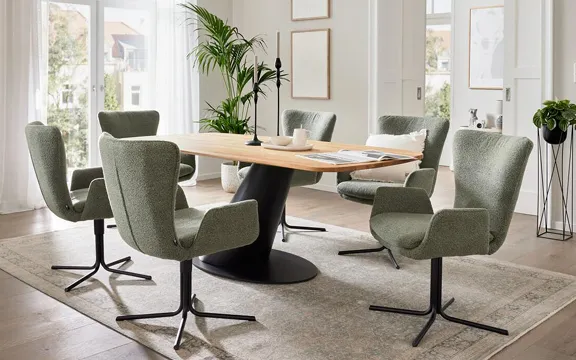 Helles Esszimmer mit modernem Esstisch und sechs grünen Stühlen. Große Fenster lassen viel Licht herein, während Pflanzen das Ambiente betonen.