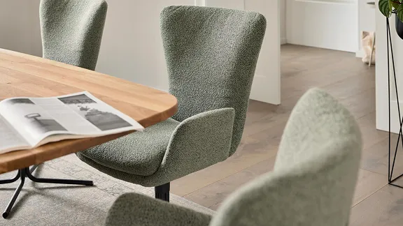 Moderner Essbereich mit salbeigrünen Polsterstühlen und einem Holztisch. Eine aufgeschlagene Zeitschrift liegt auf dem Tisch und trägt zur gemütlichen Atmosphäre bei.
